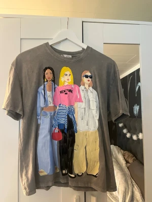 Grå t-shirt med färgglatt tryck - Cool grå t-shirt med ett stort färgglatt tryck av tre personer på framsidan. T-shirten har rund hals och korta ärmar. Materialet känns som mjuk bomull och passformen är normal. Perfekt för dig som gillar streetstyle och unika prints.