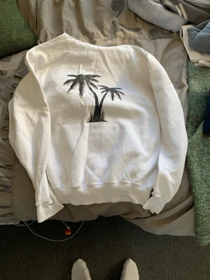 Vit sweatshirt med palmtryck Fruit of the Loom - Vit sweatshirt från Fruit of the Loom i storlek S. Tröjan har ett svart tryck med två palmer på ryggen och är gjord i mjukt bomullsmaterial. Klassisk passform utan huva och med ribbade muddar vid ärmslut och nederkant. Perfekt för en chill och avslappnad stil.