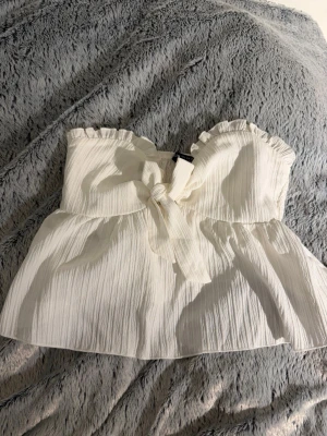 Vit axelbandslös topp med knyt - Söt vit axelbandslös topp med volangkant upptill och knyt framtill. Tillverkad i ett lätt, strukturerat tyg som ger en luftig känsla. Perfekt för varma dagar och enkel att styla med jeans eller kjol.