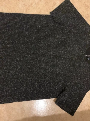 Glittrig svart topp från Pieces - Snygg svart topp från Pieces med glittrigt material och korta ärmar. Perfekt för dig som vill sticka ut lite extra. Enkel rak passform och mjukt stretchigt tyg som känns bekvämt hela dagen.