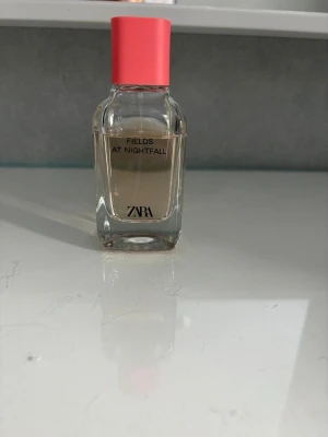 ZARA Fields at Nightfall parfym - ZARA Fields at Nightfall är en parfym i en stilren, transparent glasflaska med en ljusrosa kork. Flaskan har raka former och enkel design med svart text. Doften är elegant och flaskan känns lyxig att ha framme på hyllan.