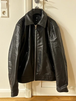 Svart skinnjacka från H&M - Snygg svart skinnjacka från H&M med klassisk krage och dragkedja framtill. Jackan har två snedställda fickor med dragkedja och en rak passform. Använd bara en gång. Nypris ca 800