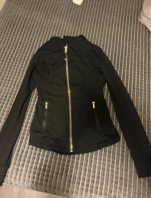 Svart vindjacka från Lululemon - Snygg svart vindjacka från Lululemon med figurnära passform och hög krage. Jackan har hel dragkedja framtill, två fickor med dragkedja och diskret logga på ryggen. Tillverkad i ett lätt och stretchigt material som passar perfekt till vardags eller träning.