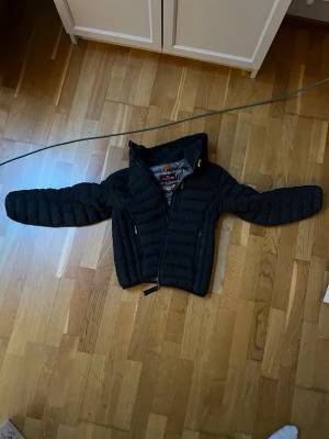 Svart dunjacka från Parajumpers - Säljer en svart dunjacka från Parajumpers med huva och dragkedja framtill. Jackan har två sidofickor med dragkedja och en patch på ärmen. Klassisk quiltad design och gul detalj vid halsen. Perfekt för kalla vinterdagar.