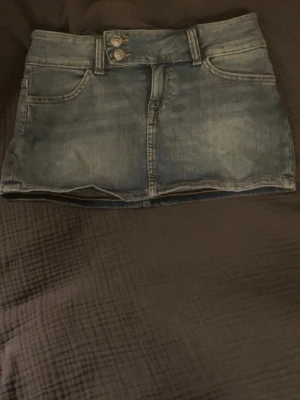 Kort jeanskjol i blå denim - Säljer en klassisk blå jeanskjol i kort modell med fickor både fram och bak. Kjolen har bälteshällor och en rak passform som ger en avslappnad look. Perfekt att matcha med en t-shirt eller hoodie för en cool vardagsstil.