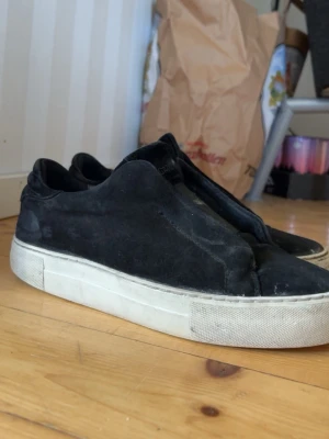 Svarta slip-on sneakers mocka - Svarta slip-on sneakers i mocka med tjock vit sula. Skorna har en minimalistisk design utan snörning och en låg profil som gör dem enkla att ta på. Perfekta för en clean och avslappnad stil.