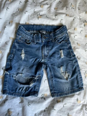 Blå slitna jeansshorts med hål Barn st 122 - Säljer ett par blå jeansshorts från H&M med slitna detaljer och hål på framsidan. Klassisk femficksmodell med knapp och dragkedja. Perfekta för en avslappnad och trendig look. Materialet är bomull med en skön denimkänsla. Barn med storlek 122
