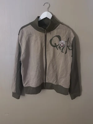 1970 Quiksilver Beige vindjacka  - Snygg beige vindjacka från Quiksilver med gröna detaljer och stort tryck på bröstet. Jackan har hel dragkedja framtill, hög krage och muddar vid ärmslut och nederkant. Materialet är lätt och passar perfekt för blåsiga dagar.