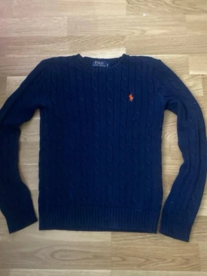 Mörkblå stickad tröja Polo Ralph Lauren - Klassisk mörkblå stickad tröja från Polo Ralph Lauren med kabelstickat mönster och den ikoniska orange loggan broderad på bröstet. Tröjan har rund halsringning och långa ärmar, perfekt för lager på lager under höst och vinter.