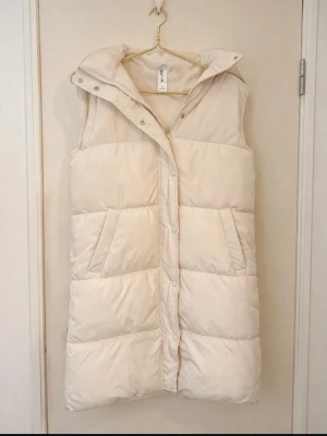 Ljusbeige lång dunväst  - Stilren ljusbeige lång dunvästmed hög krage och stora fickor framtill. Västen har dragkedja och tryckknappar samt en quiltad design som ger en puffig look. Perfekt för lager-på-lager och trendig streetstyle.