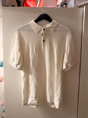 Vit stickad polotopp med knappar - Klassisk vit stickad polotopp med korta ärmar och tre knappar framtill. Toppen har en mjuk, strukturerad yta och en avslappnad passform. Perfekt till jeans eller kjol för en clean look.