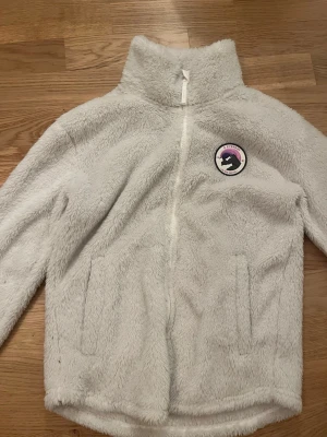 Vit teddyjacka med patch från Everest - Mysig vit teddyjacka med hög krage och dragkedja framtill. Jackan har två fickor och en rund patch med motiv i lila och svart på bröstet. Perfekt för kalla dagar och ger en trendig look. I storlek 158/164. Pris kan diskuteras.