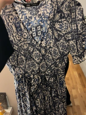 Blåvit mönstrad kortklänning Zara - Säljer en blåvit kortklänning från Zara med v-ringning och korta ärmar. Klänningen har ett unikt blommigt och geometriskt mönster, markerad midja och lätt utställd kjol. Perfekt för dig som gillar en luftig och trendig stil.