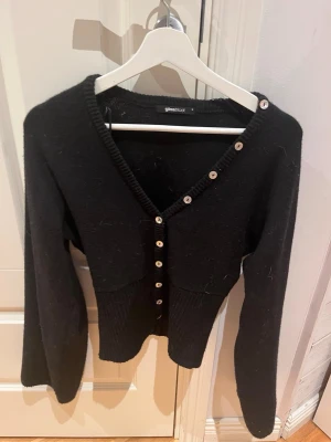 Svart kofta med knappar från Gina Tricot - Snygg svart kofta från Gina Tricot med v-ringning och dekorativa vita knappar längs hela framsidan. Koftan har lång ärm och ribbad midja för en smickrande passform. Perfekt att styla över ett linne eller t-shirt.