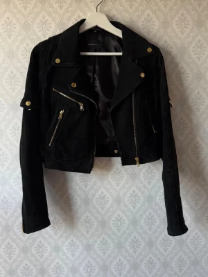 Mockajacka Rock n Blue - Snygg svart jacka i mocka från Rock'n Blue med guldiga detaljer och dragkedjor. Croppad modell med bred krage och flera fickor framtill. Jackan har lång ärm. Perfekt för dig som vill ha en statement piece i garderoben. Knappt använd. Rökfritt hem.