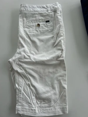 Vita shorts från Polo Ralph Lauren - Klassiska vita shorts från Polo Ralph Lauren med rak passform och bakficka med knapp. Tillverkade i mjuk bomull och har bälteshällor i midjan. Perfekta för varma dagar och enkel att matcha med olika stilar.
