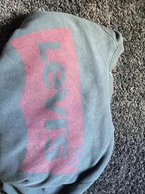 Grå Levi's hoodie med stor logga - Snygg grå hoodie från Levi's med stor rosa logga framtill. Klassisk modell med lång ärm och rund halsringning. Tillverkad i mjukt bomullsmaterial som är skönt mot huden. Perfekt för en avslappnad streetwear-look.