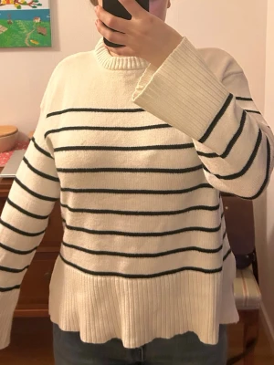 Randig vit stickad tröja - Snygg beige stickad tröja med svarta horisontella ränder. Tröjan har rund halsringning och långa ärmar med samma randiga mönster och slits på båda sidorna.. Perfekt för dig som gillar klassisk och enkel stil. Materialet känns mjukt och bekvämt. Från lager  157 