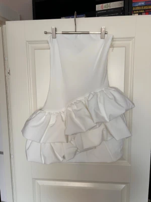 Vit kort klänning med volanger - Supersnygg vit kort klänning med asymmetriska volanger och ärmlös design. Klänningen har en modern och trendig look, perfekt för dig som vill sticka ut. Tillverkad i ett slätt tyg som ger en lyxig känsla.