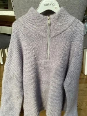 Ljuslila stickad tröja med halvzip - Mysig ljuslila stickad tröja med hög krage och halvzip framtill. Tröjan har breda ribbstickade muddar och en avslappnad passform. Perfekt för kalla dagar och enkel att styla till olika outfits. Köparen står för frakt.