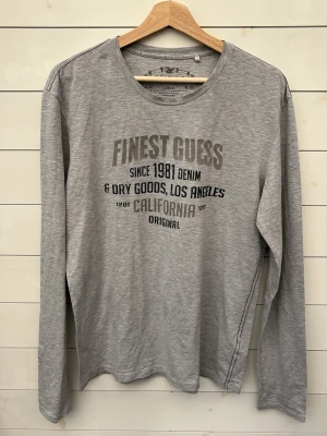 Grå långärmad topp från Guess - Snygg grå långärmad topp från Guess med tryck på bröstet i svart text. Tillverkad i mjuk bomull som känns skön mot huden. Perfekt för en avslappnad och cool stil med rund halsringning och normal passform. Unisex 