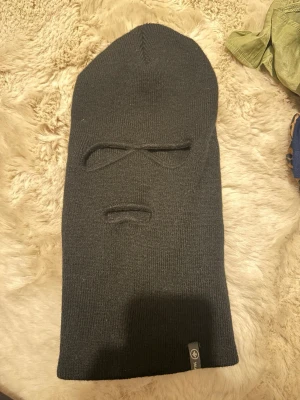 Svart balaclava i ribbstickad bomull - Säljer en svart ribbstickad balaclava med två öppningar för ögon och en för mun. Tillverkad i mjuk bomull och har en liten tygpatch med logga längst ner. Perfekt för kalla dagar när du vill hålla dig varm och anonym.