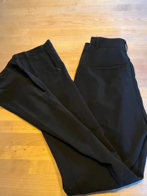 Svarta kostymbyxor med raka ben - Snygga svarta kostymbyxor med raka ben och diskreta sidofickor. Byxorna har hällor för bälte och klassisk dragkedja framtill. Perfekta för en stilren och clean look. Materialet ser ut att vara en mjuk och följsam mix, troligen polyester eller bomull.