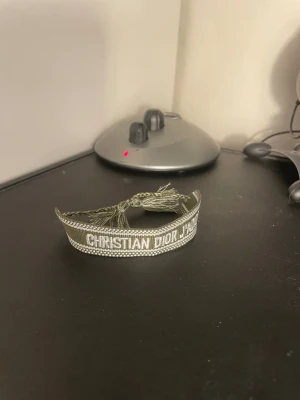 Christian Dior J'Adior armband - Snyggt armband från Christian Dior med texten 'J'Adior' broderat i vitt på en olivgrön bakgrund. Armbandet är gjort i textil och har dekorativa tofsar i ändarna för en trendig look. Perfekt accessoar för att lyfta din outfit.