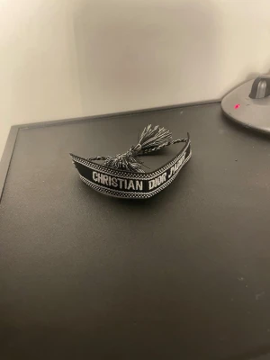 Christian Dior svart tygarmband - Snyggt svart tygarmband från Christian Dior med texten 'CHRISTIAN DIOR J'ADIOR' i vitt. Armbandet har dekorativa tofsar i ändarna och ett stilrent, vävt mönster längs kanterna. Perfekt accessoar för att lyfta din outfit.