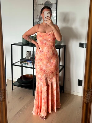 Adoore klänning - mer som strl 38 - Säljer en lång, figurnära klänning med smala axelband och blommigt mönster i orange, rosa och ljusa toner. Klänningen har en mjukt draperad urringning och är tillverkad i ett lätt, följsamt material som passar perfekt till sommaren.