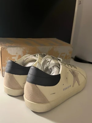 Golden Goose vita sneakers med stjärna - Golden Goose sneakers i vitt läder med beige mockadetaljer och grå stjärna på sidan. Skorna har svart hälparti, vita skosnören och en klassisk låg silhuett. Snygg vintage-look och robust gummisula. Perfekta för dig som gillar streetstyle och unika detaljer.