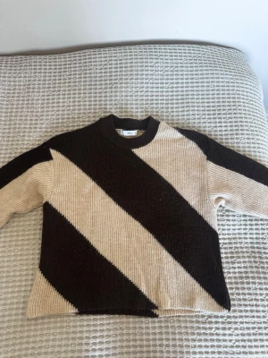 Beige och brun stickad tröja Mango - Säljer en cool stickad tröja från Mango med breda diagonala ränder i beige och mörkbrunt. Tröjan har rund hals och långa ärmar, perfekt för att hålla sig varm. Materialet är mjukt och grovt stickat, vilket ger en skön känsla.