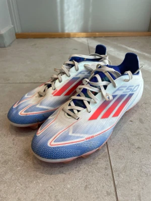 Adidas F50 pro blåvita fotbollsskor MG - Säljer ett par Adidas F50 PRO fotbollsskor i blått, vitt och rött med klassiska tre ränder på sidan. Skorna har snörning, låg profil och dobbar MG (multiground) i rosa  för bra grepp på både naturgräs och konstgräs. Ovandelen är i syntetmaterial och designen är riktigt sportig och snabb. Skorna är andvänd bara några fåtal gånger på naturgräs. Du får en unisport påse med på köpet. Skorna köpt i originalpris 1799kr