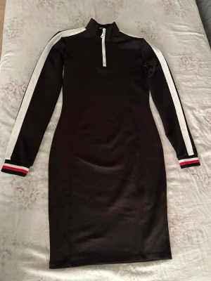 Svart sportig klänning med vita ränder - Svart långärmad klänning med vita ränder längs ärmarna och en vit half zip-dragkedja framtill. Klänningen har en sportig look med ribbade muddar i svart, vitt och rött. Figurnära passform och hög krage. Perfekt för en trendig och avslappnad stil.