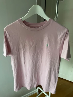 Rosa t-shirt från Polo Ralph Lauren strl 14-16 - Säljer en ljusrosa t-shirt från Polo Ralph Lauren med klassisk passform och korta ärmar. T-shirten har en liten broderad logga på bröstet och är gjord i mjuk bomull. Perfekt till jeans eller shorts för en clean och enkel look. Junior 14-16