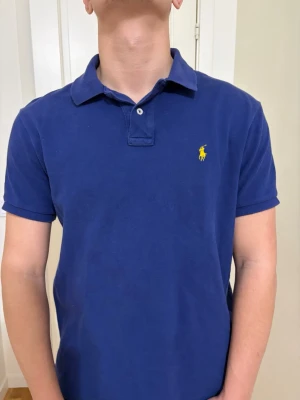 Marinblå pikétröja från Ralph Lauren - Klassisk marinblå pikétröja från Ralph Lauren med gul broderad logga på bröstet. Tröjan har korta ärmar, krage och knäppning med två knappar. Perfekt till jeans eller shorts för en clean och stilren look. L men passar som M