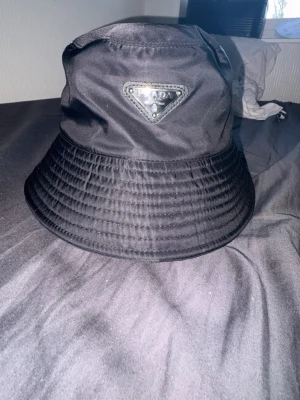 Svart bucket hat från Prada - Snygg svart bucket hat från Prada i glansigt nylonmaterial. Klassisk form med bred, nedåtlutande brätte och ikoniska metalltriangeln med logga framtill. Perfekt accessoar för en trendig och avslappnad stil.