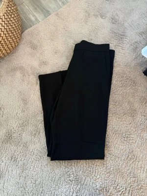 Svarta kostymbyxor med raka ben - Snygga svarta kostymbyxor med hög midja och raka ben. Klassisk design med pressveck framtill och diskreta fickor i sidorna. Perfekta för en clean och stilren look. Materialet ser ut att vara en mjuk och följsam mix, troligtvis polyester eller viskos.