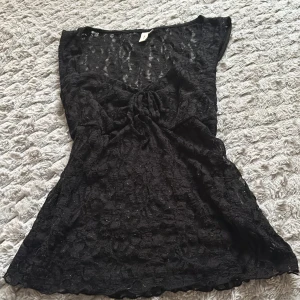 Svart spets strandtunika med knyt - Snygg svart strandtunika i genombruten spets med knyt framtill. Tunikan har korta ärmar och ett lätt, luftigt material som passar perfekt över bikinin. Transparent design ger en trendig look på stranden.