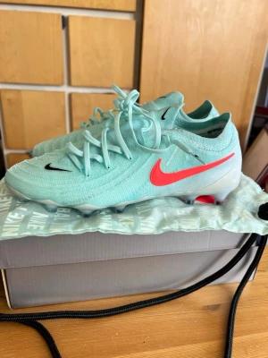 Nike turkosa fotbollsskor med röd swoosh - Snygga fotbollsskor från Nike i en fräsch turkos färg med en iögonfallande röd swoosh på sidan och svart Nike-logga på tån. Skorna har snörning och är tillverkade i ett lätt syntetmaterial som ger bra känsla på planen. Perfekta för dig som vill sticka ut på matchen. Använda på 1 träning. De är för små för mig som brukar använda storlek 42. Dyraste modellen, allt medkommer