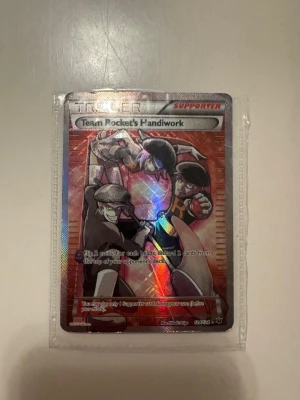 Pokémon Team Rocket's Handiwork kort - Säljer ett Team Rocket's Handiwork supporter-kort från Pokémon TCG. Kortet är holografiskt med färgstark design och motiv av Team Rocket. Perfekt till samlingen eller för dig som gillar att spela Pokémonkort. Kortet är tillverkat i laminerad papp och plast.