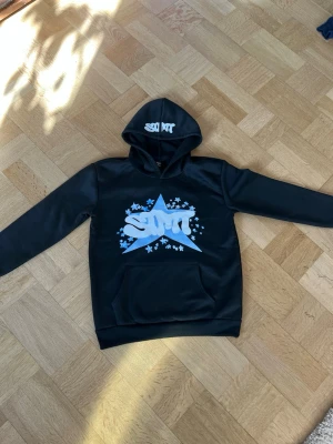 Svart hoodie med blå stjärna och STMT - Snygg svart hoodie från SMT med stor blå stjärna och vit SMT-logga på bröstet. Hoodien har huva med tryckt SMT-text och en klassisk magficka. Materialet ser ut att vara mjukt och bekvämt, perfekt för en avslappnad streetwear-look. Ásian S