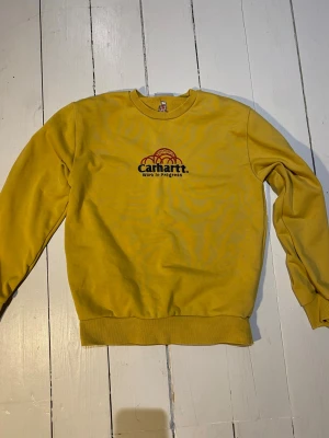 Gul sweatshirt från Carhartt WIP - Gul sweatshirt från Carhartt Work In Progress med tryckt logga och text på bröstet. Tröjan har rund halsringning, ribbade muddar vid ärmslut och nederkant. Perfekt för en avslappnad streetwear-look.