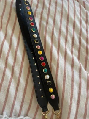 Gabrielle by P axelrem med nitar 105cm - 105cm. Snygg svart axelrem i konstläder från Gabrielle by P, dekorerad med färgglada runda nitar i gult, rosa, rött, grönt, vitt och svart. Remmen har guldfärgade metalldetaljer och karbinhakar för enkel fastsättning på väska.