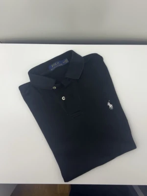 Svart pikétröja från Polo Ralph Lauren - Klassisk svart pikétröja från Polo Ralph Lauren med vit broderad logga på bröstet. Tröjan har korta ärmar, krage och två knappar framtill. Tillverkad i mjuk bomull som känns skön mot huden. Perfekt till jeans eller shorts.