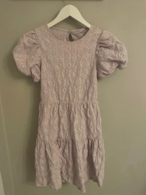 Ljuslila klänning med puffärm - Supersöt ljuslila klänning med broderade blommor och puffärmar. Klänningen har volang nertill och öppen rygg med knappstängning upptill. Perfekt för vår och sommar när du vill känna dig extra fin.