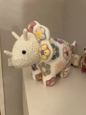 Virkad dinosaurie i pastellfärger - Unik handvirkad dinosaurie i vitt med detaljer i pastellfärger som lila, rosa, gul, blå och grön. Mjuk och fantasifull design med två horn och svans, perfekt som dekoration eller gosedjur. Tillverkad i garn med virkade mormorsrutor.