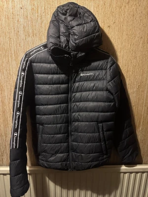 Svart pufferjacka från Champion - Svart pufferjacka från Champion med huva och dragkedja. Jackan har vita Champion-loggor längs ärmarna och en broderad logga på bröstet. Quiltad design och innerficka med mesh. Perfekt för kalla dagar och riktigt snygg streetstil.