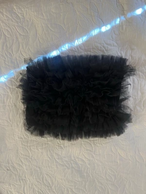 Svart tubtopp med volanger - Säljer en svart tubtopp med flera lager volanger i tyll som ger en riktigt statement look. Perfekt för dig som vill sticka ut och älskar trendiga detaljer. Toppen är axelbandslös och har en croppad passform.