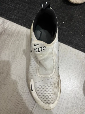 Nike Air Max 270 vita sneakers - Nike Air Max 270 sneakers i vitt med svarta detaljer och stor synlig luftbubbla i hälen. Ovandelen är i mesh och syntetmaterial, slip-on design utan klassiska snören och tydliga Nike-loggor på sidan och tungan. Sportig och modern look.